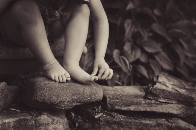 A black and white close up of a toddler's hands and feet by a creek. Jesus was born a baby on earth so he could experience all that humans do, even though he is fully God.