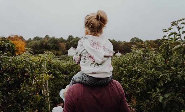 A young girl sits on her father's shoulders as they walk around a field of fruit bushes. there is a farm house in the distance. God protects His children and loves them just as a father on Earth would. That is why He became a man, to save them from a life in eternity without Him.