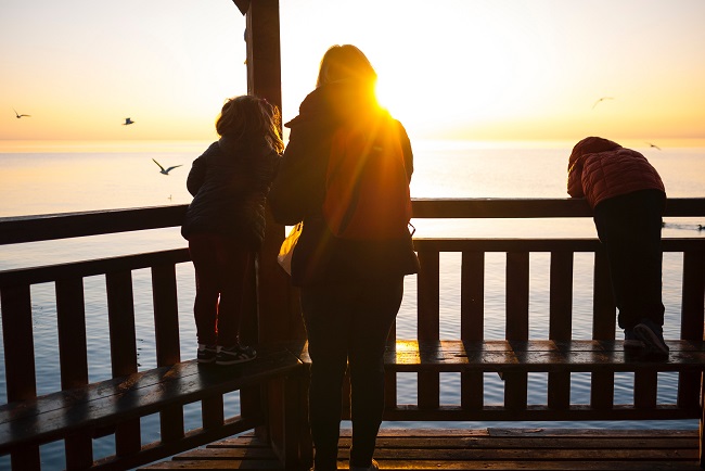 A mom and her children stand together ona balcony was the sun sets over the ocean in front of them. The sun hits her shoulder and we see the family in silhouette. Heaven came down to earth to save God's children, in the form of a man.