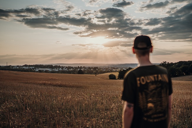 A young man stands with his back to the camera, looking off towards the sun that casts brilliant rays across the earth, piercing through low clouds. Jesus came to earth and was taught by many, including God and Mary and Joseph.