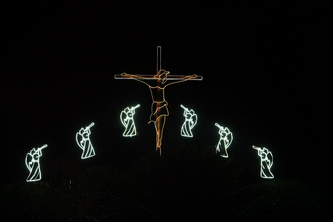 Lights against a black background depict the crucifiction of Jesus, with angels on either side of him. Why did Jesus come to earth? Read this article to find out!