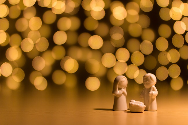 A small stone nativity sits on a table in front of glittering lights in the background. Jesus came to earth to save us!