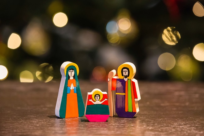A colorful nativity set sits upon a table with Christmas lights behind them in the distance. Jesus came into the world as a man at exactly the right time, this blog post explains the circumstances.