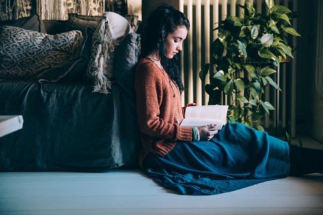 A young woman in a sweater and skirt, sits on the floor leaning up against a sofa. She is reading her Bible that is great for new believers!