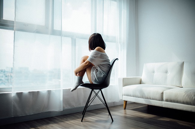 A yound woman sits on a black chair, hugging her knees to her chest. She looks out of a large wall-length window with sheer white curtains across it. God is here for you in your grief, reach out to Him for help.