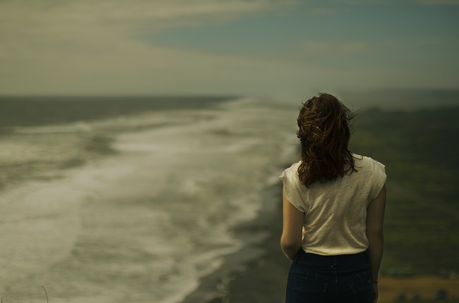 A woman walks along a beach. The lighting is dark and moody, with an unfriendly gray sky. God can help you in your grief with His 12 steps to Recovery.