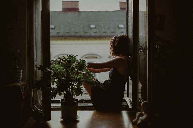 A woman sits on a windowsill and looks out of the open window with her legs tucked up and arms resting on her knees. Give God your struggles and He will answer with peace. 