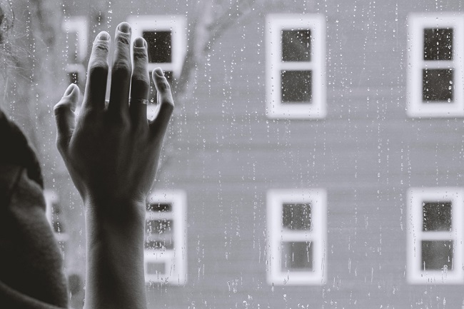 A gray-scale photo of a woman's hand on a glass pane, rain falls outside and the window looks out onto a building with lots of little windows. God can help you with recovery from your grief.