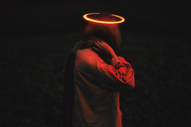 A young woman stands in a dark room with a red neon halo on her head. We can't see her face and her hand is placed by her neck. What will we all look like in Heaven? Probably not too dissimilar to how we look now!