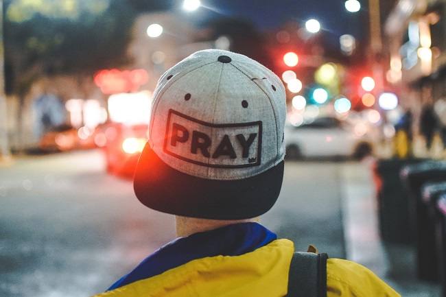 A young man is walking down the street with his cap on backwards, it says 'PRAY' on his hat.