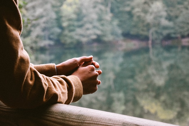 A man's hands can be seen in prayer, leaning on a balcony over-looking a beautiful river and tress on the other side.