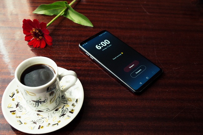 A smart phone sits on a dark wooden table with a full coffee cup and a flower laying next to it.