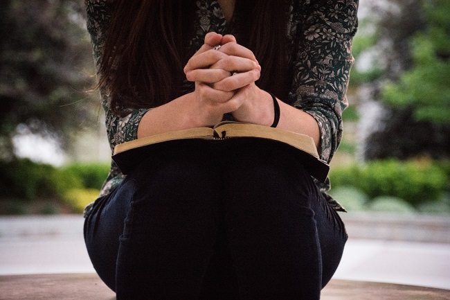 A woman sits with her hands clasped in prayer over her bible that lays on her lap.