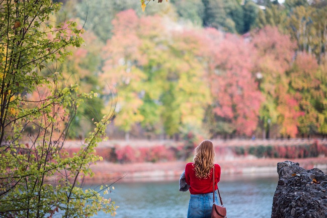 A woman stands by a lake surrounded by colorful early fall trees. Silence is an important part of prayer.