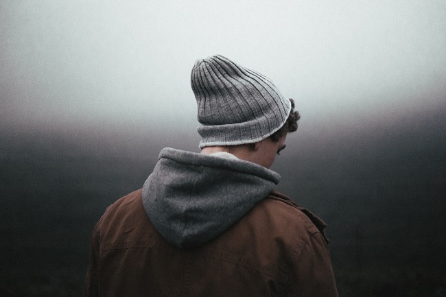 A young man in a jacket and beanie hat, stands with his back to the camera. in the background all we see is light and dark gray tones.