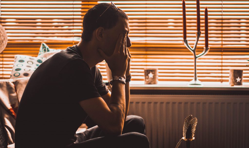 A sad man sits praying in the dark next to a window.