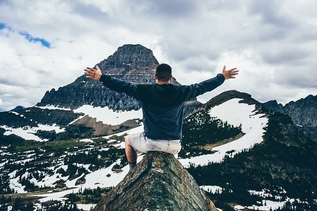 A joyful man sits atop a snowy mountain peak with a large mountain ahead of him.