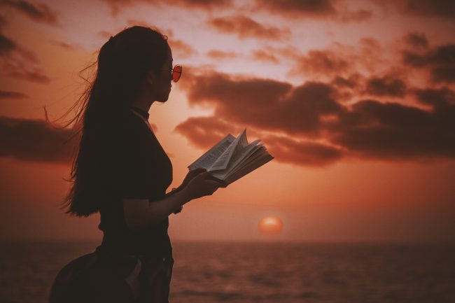 Young woman by the sea with a Bible as the sun sets. Encouraging quiet time and Bible reading.