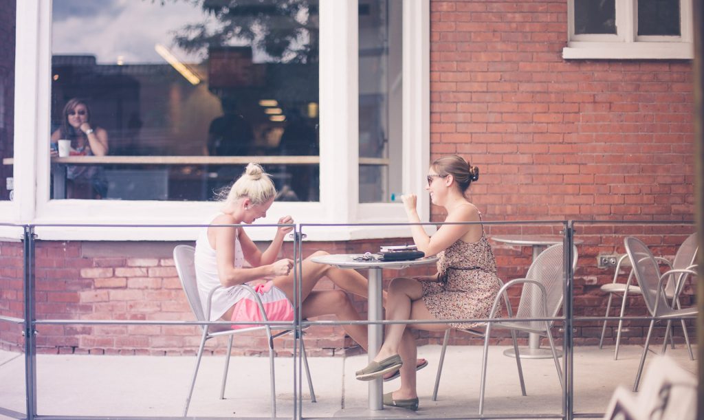 Two women sit at outside a cafe laughing, having a spiritual conversation.