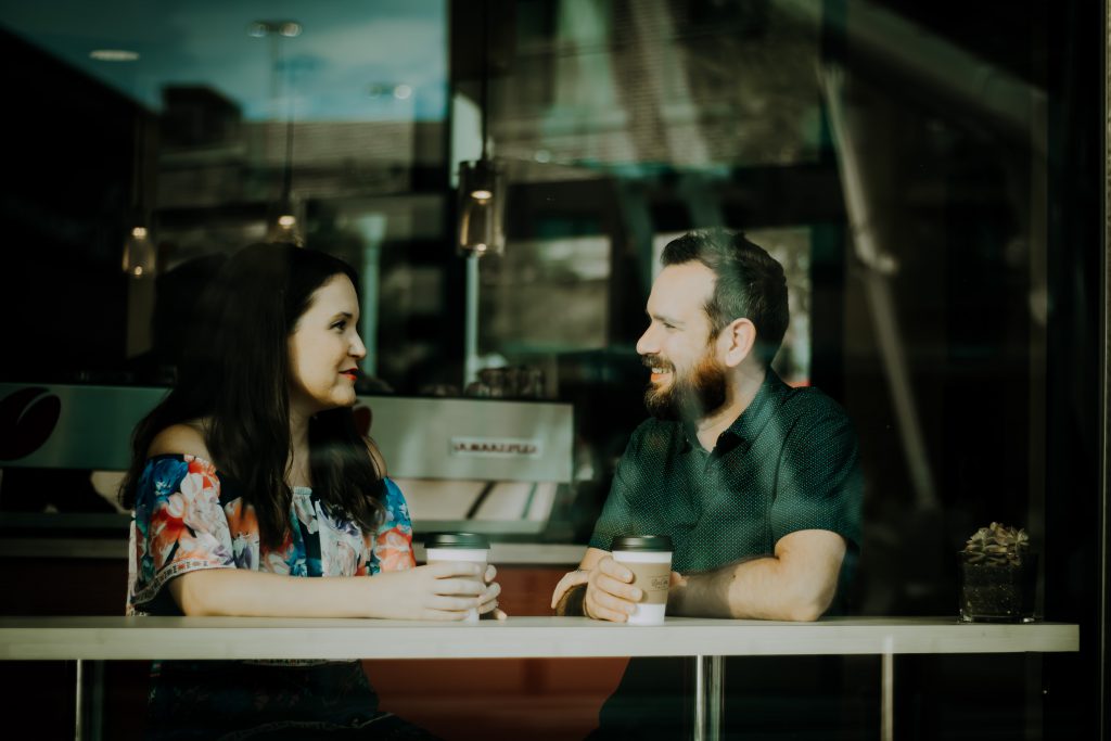 Male and female couple talk about faith in a cafe, seen through a large window.