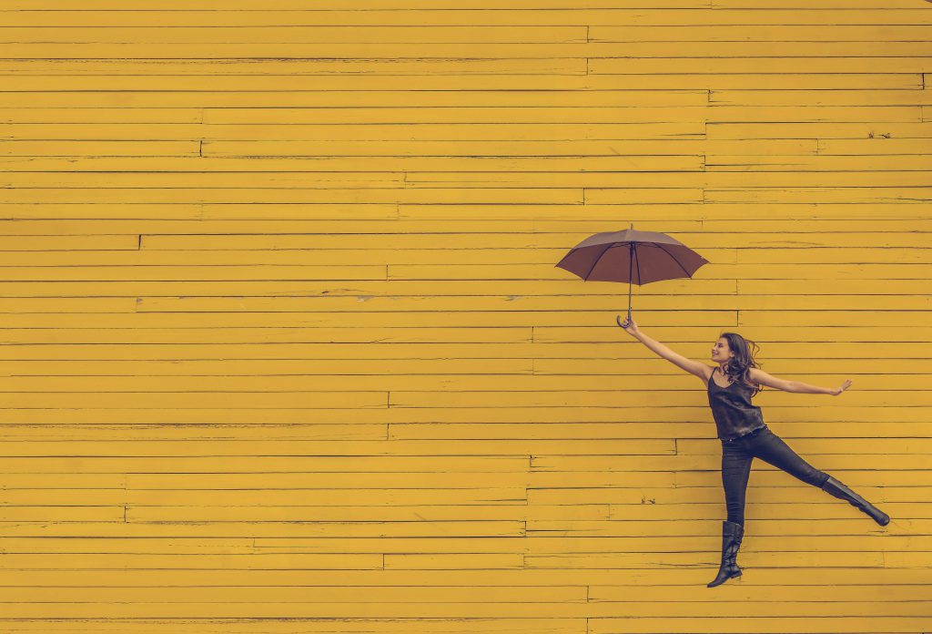 Woman jumping for joy with an open umbrella, in front of a yellow wall. best devotionals for new believers, 60 days to Happiness devotional.