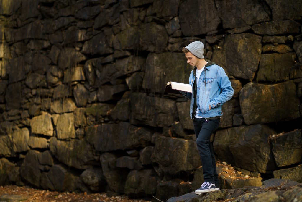 A young man with a denim jacket and beanie reads the Bible in front of an old stone wall. He loves his Bible study.