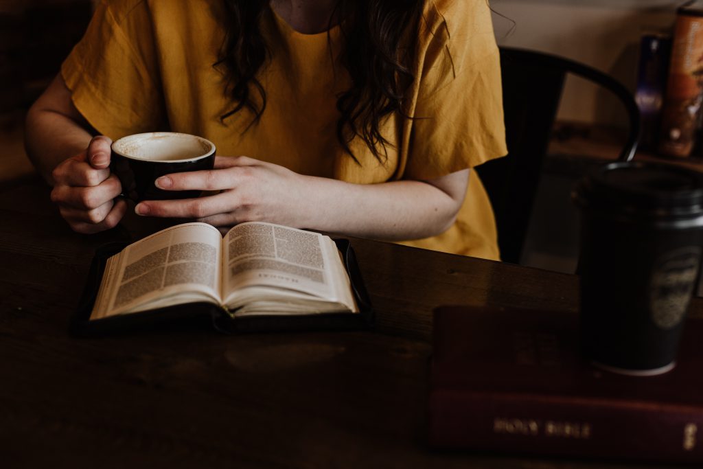 A lady in a yellow top and coffee in her hands reads her Bible study on top of a wooden table.