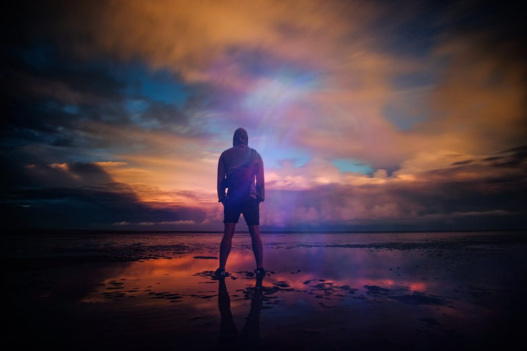 God offers hope in all situations, especially when forgiving our sin. A young man stands on a wet beach with a moody, colorful sky reflected on the still water.