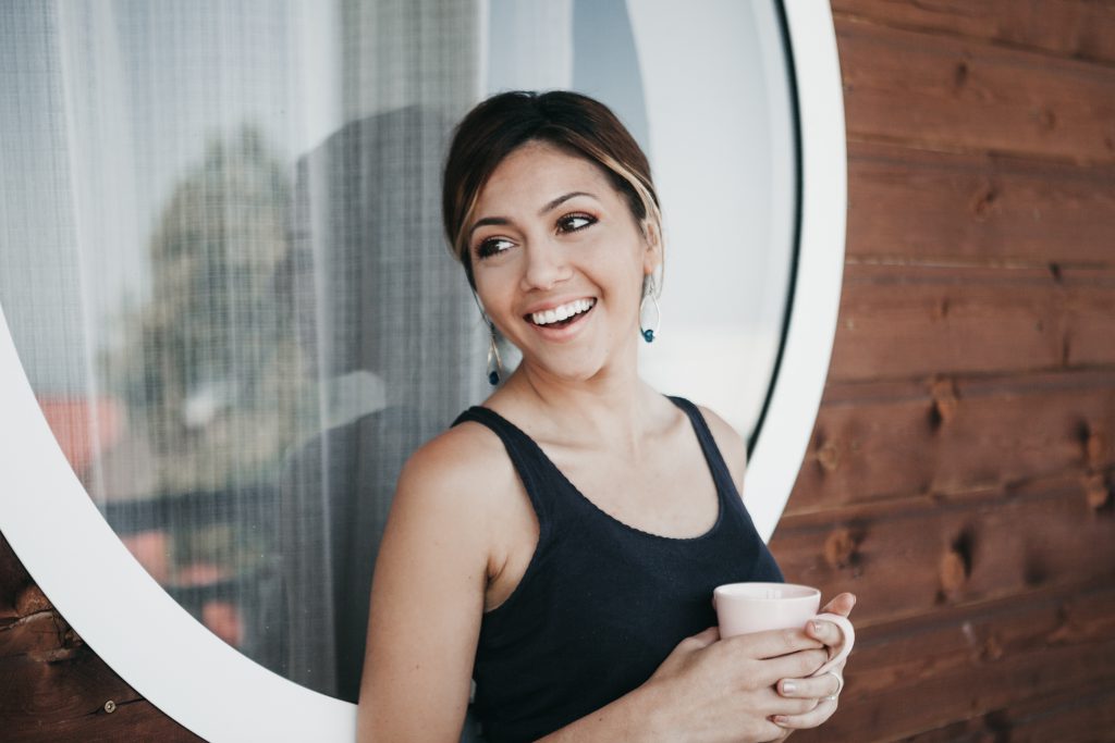 Sharing your testimony is a powerful tool for sharing the love of Jesus. Girl with coffee outside a window, smiling as she talks to an off-screen friend.