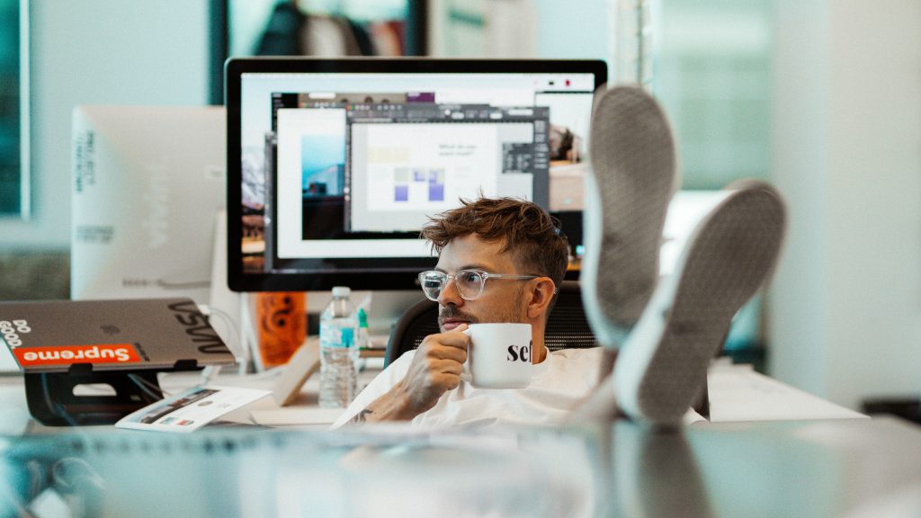 A hipster man sits with coffee in hand and his feet up on a desk, surrounded by computer equipment. Wondering how he can step away from his sinful ways.