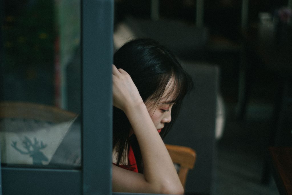 A young Asian woman looks down towards the floor in a moody shot. Thinking about how she might be strong enough to stop sinning.