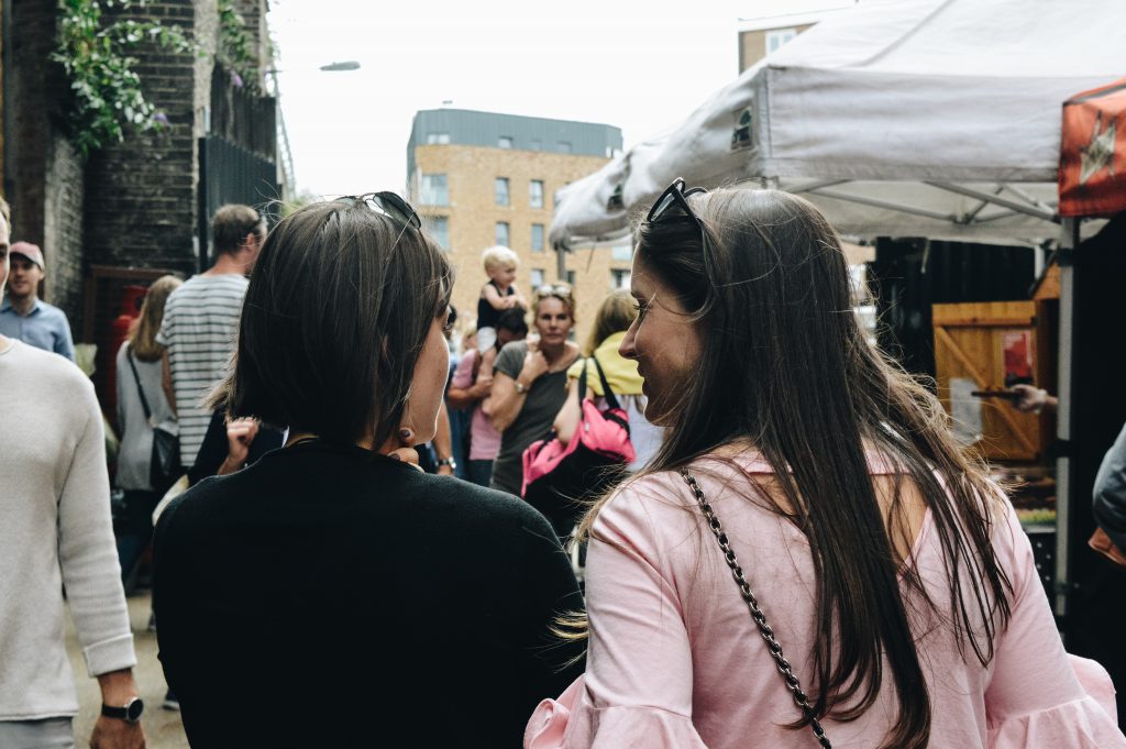 Two women walk arm in arm through a street market, we see them from the back and can see that they are talking happily about Jesus.