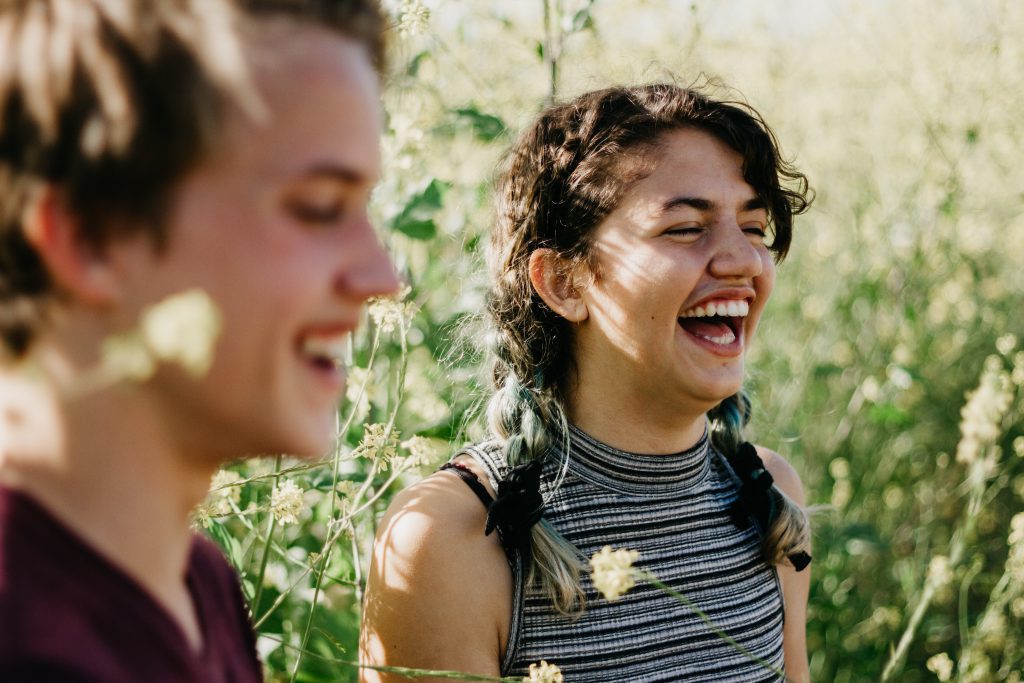 A teenage girl and boy stand in a sunny meadow laughing. They know the jot there is in talking about, and sharing, Jesus with their non-believer friends.