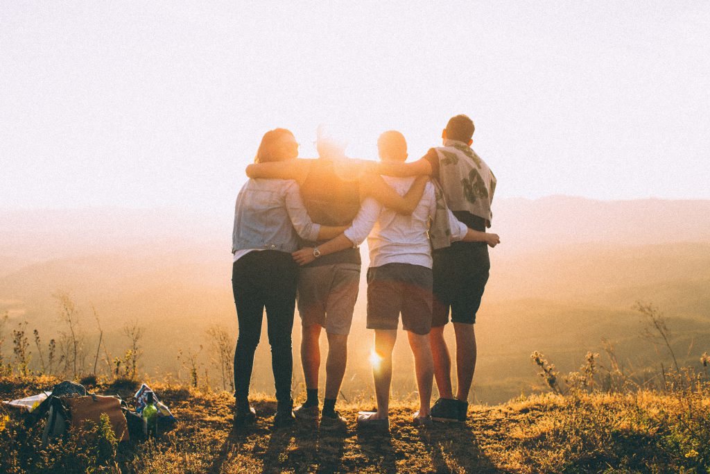Four small group friends stand together on top of a hill, watching the sunset.