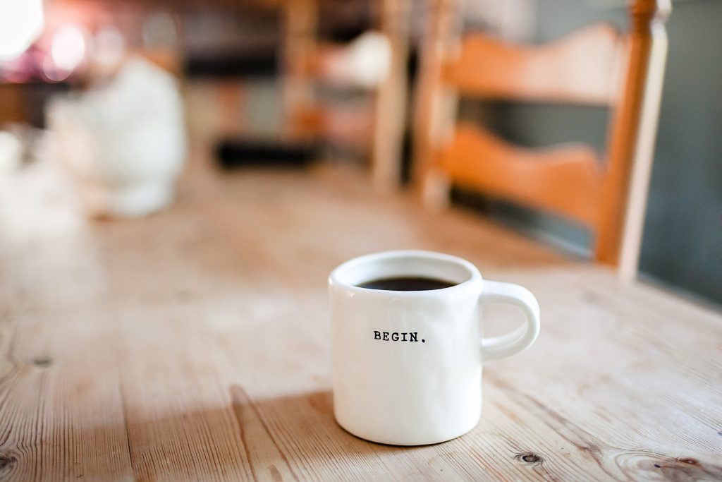 A cup of coffee sits on a long wooden table with the word 'begin' written on it. God's grace gives us a brand new start in life, so why not accept this grace and start anew in the new year?