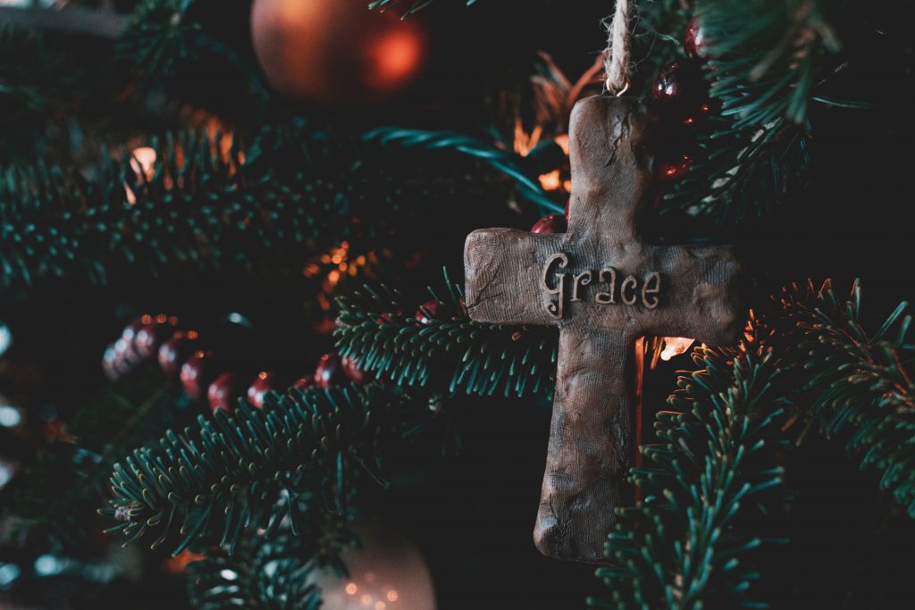 A cross with the word Grace written on it sits in a lit Christmas tree. Grace is one real gift of faith you could give this Christmas.