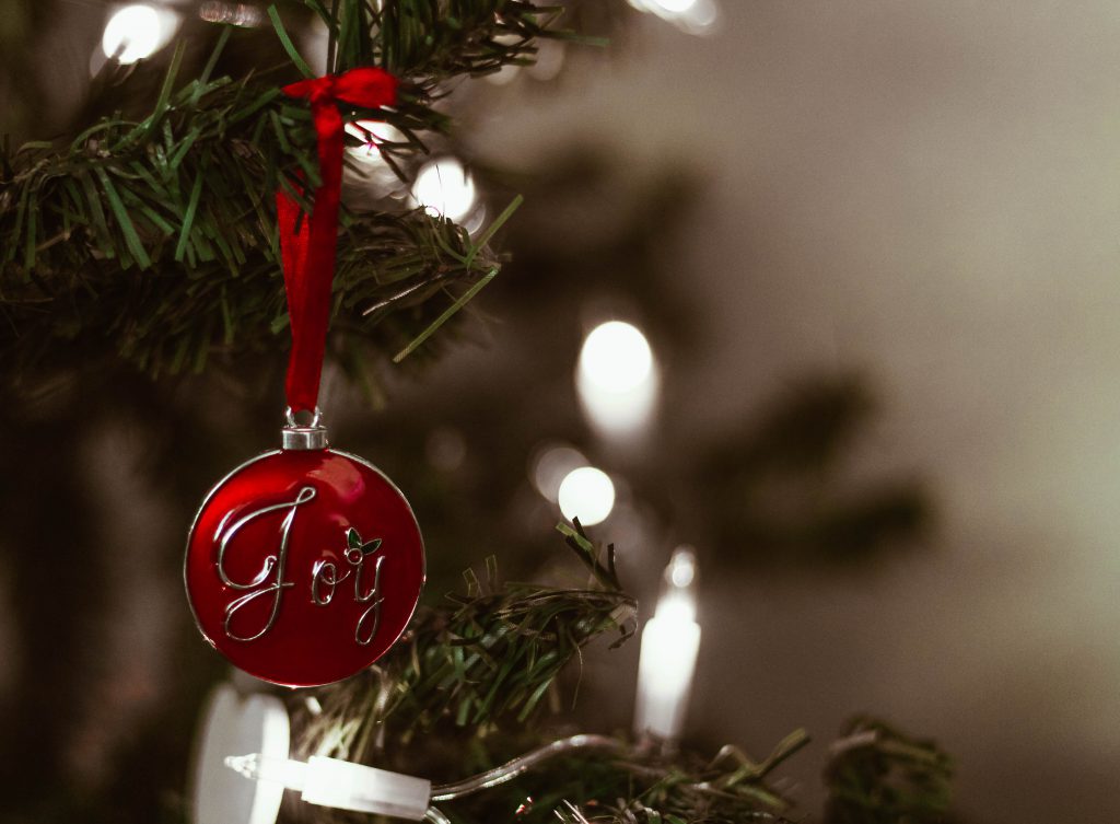 A red ornament with the word Joy written on it hangs from a Christmas tree branch with lights in the background. Joy is one real gift of faith you could consider giving this Christmas.