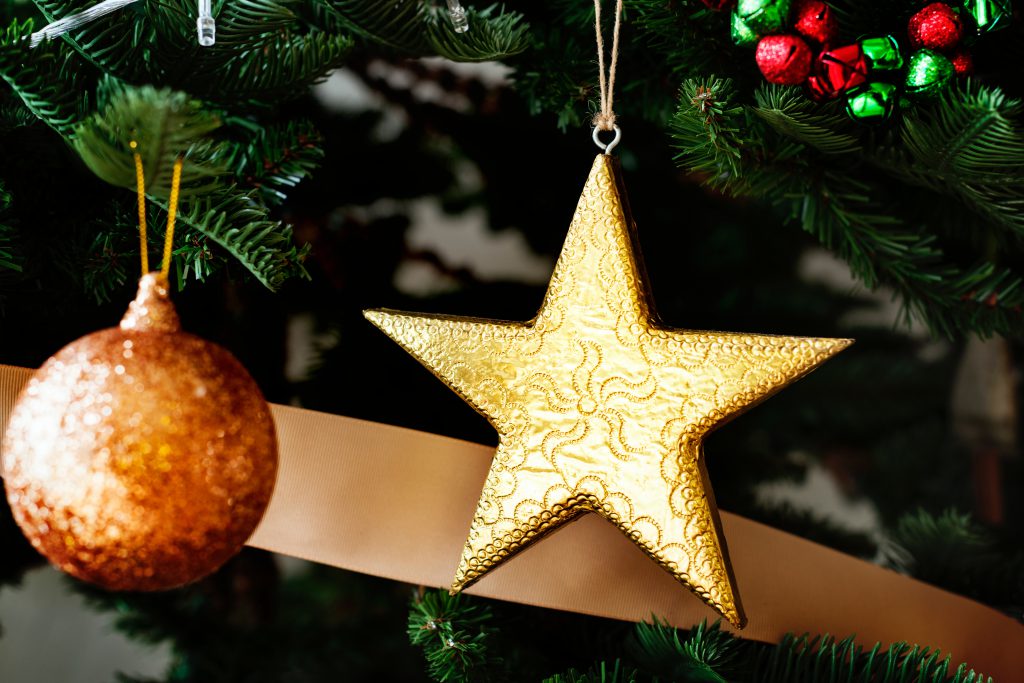 A golden star and an orange Christmas ornament hang from a decorated Christmas tree. Think and pray about which real gift of faith you could give someone this Christmas.