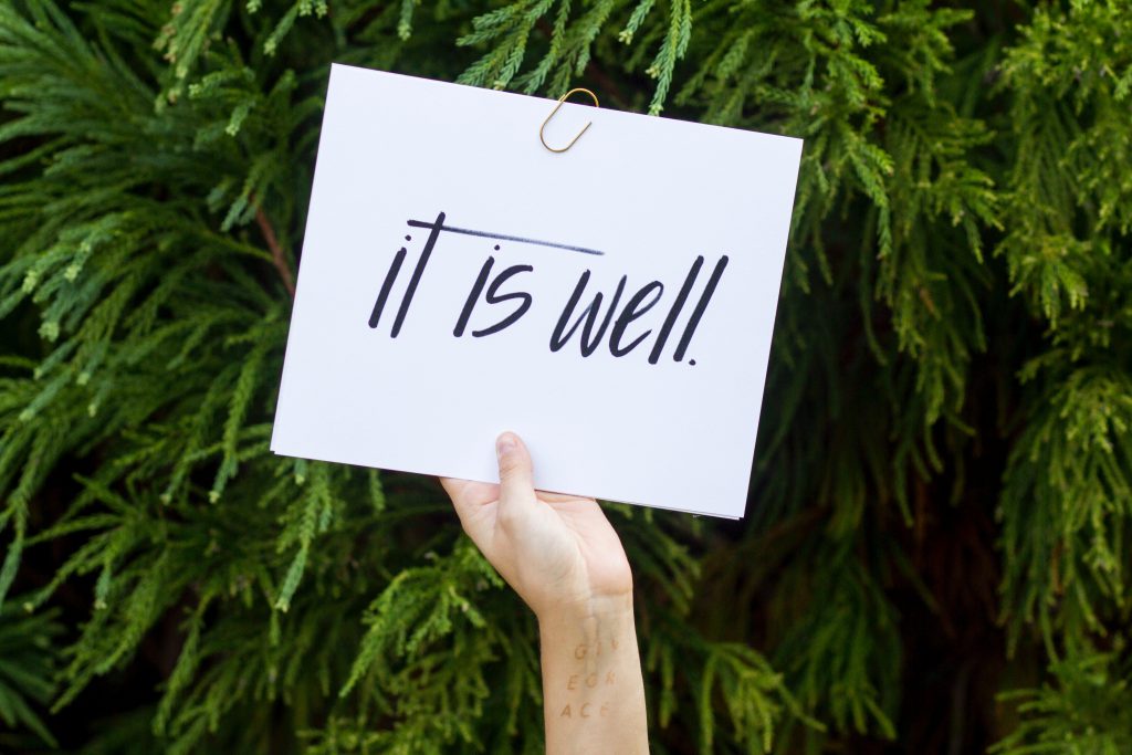 Lament can lead us back to hope. A sign is held up by a single hand in front of a green bush. The sign says 'It is well'.