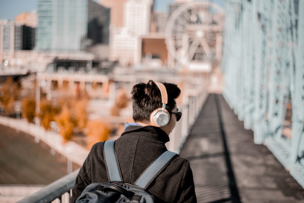 An Asian man walks across a bridge in a large city, with headphones on and listening to a podcast.