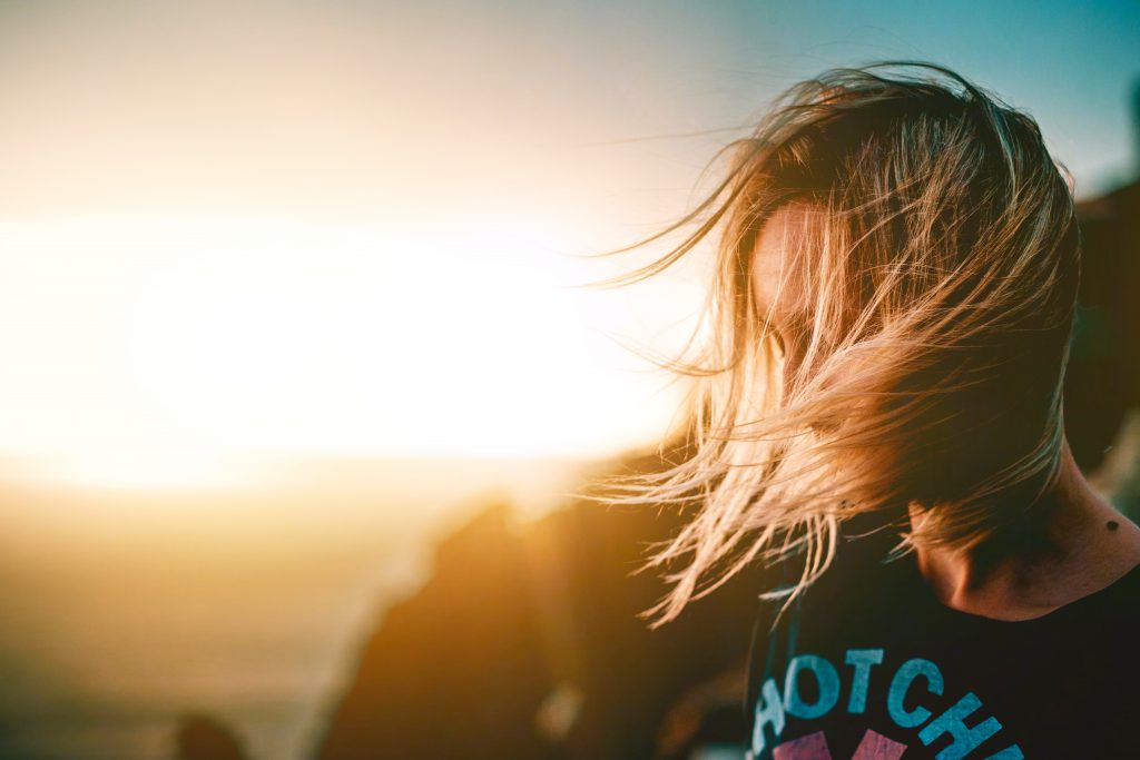 Even through our grief, in lament, there is hope in God. A blonde woman stands on a hilltop, with her hair blowing over her face in the wind. The sun sets behind her over the ocean.