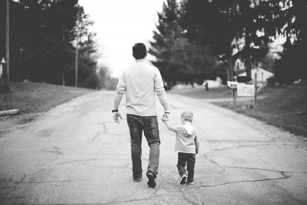 A black and white shot of a father and son holding hands as they walk down a cracked road with trees on either side of them and buildings ahead of them. Effective fathers of faith fight for their families.