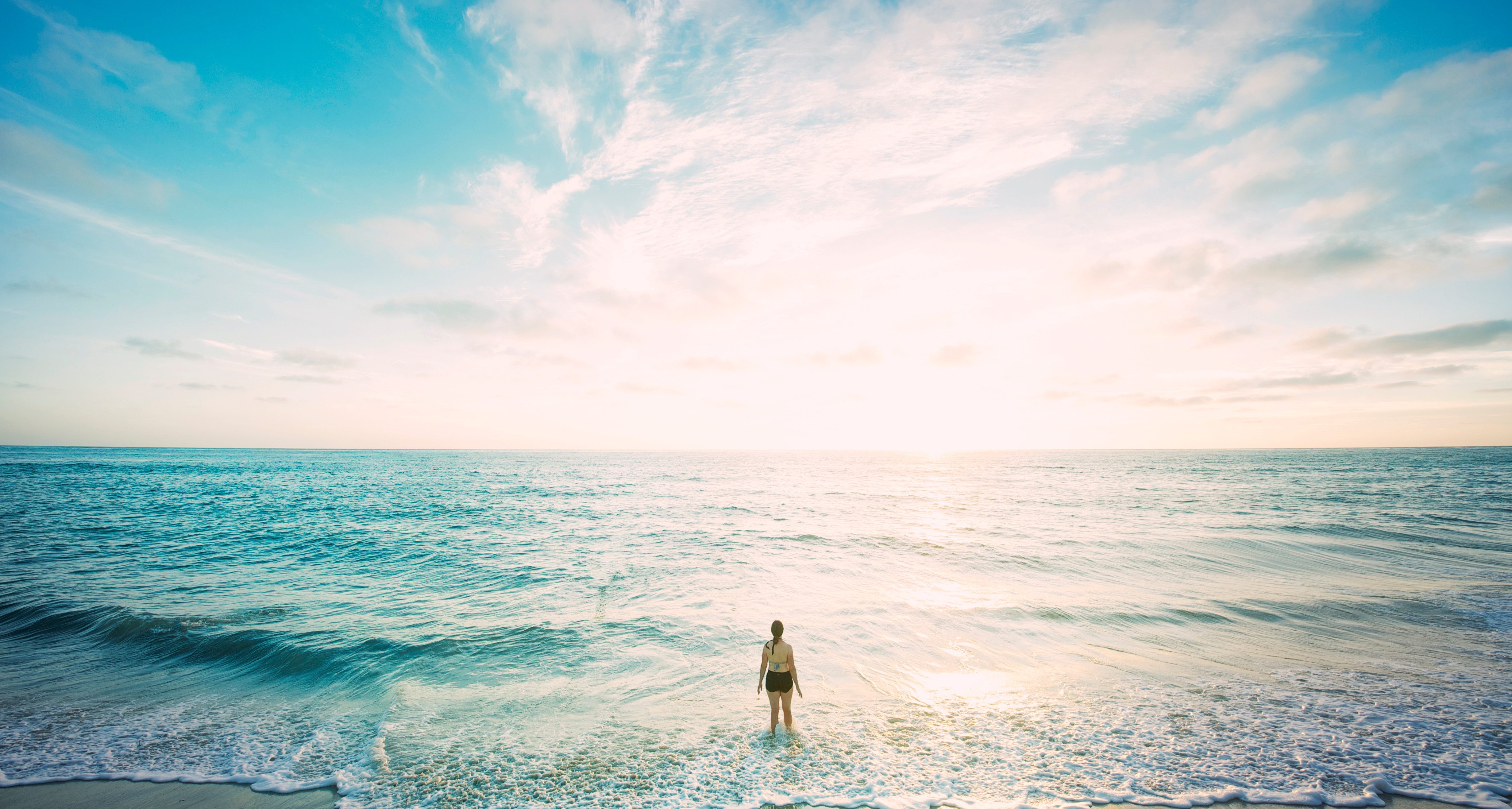 A woman in a swim suit stands within the water's edge on a beach. The sun is rising on the horizon and fills the background with light, making the beautiful blue and purple ocean look both inviting and endless. Much like God's promises, which are also inviting and endless. Here's how you can start relying on God's promises.