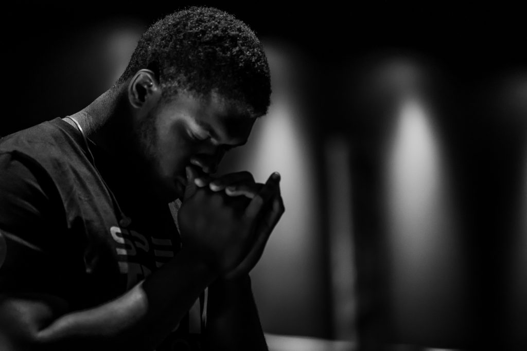 A young man clasps his hands together in front of his face, with his head bowed and sharp lights point down the walls behind him, out of focus. One of the ways in which can start to rely on God's promises is by praying for them.