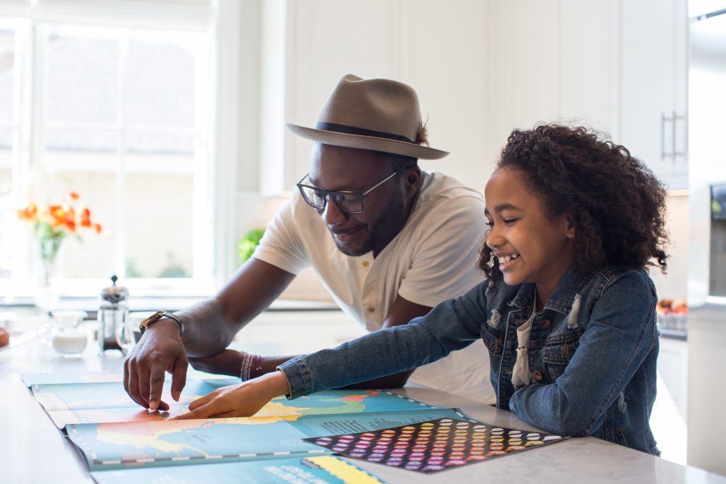 A preteen girl and her father smile as they point to a country on a colorful map. They are stood in a bright and airy white kitchen. Recognizing that God's promises cover all our needs, helps us to relax and enjoy life to the fullest. 