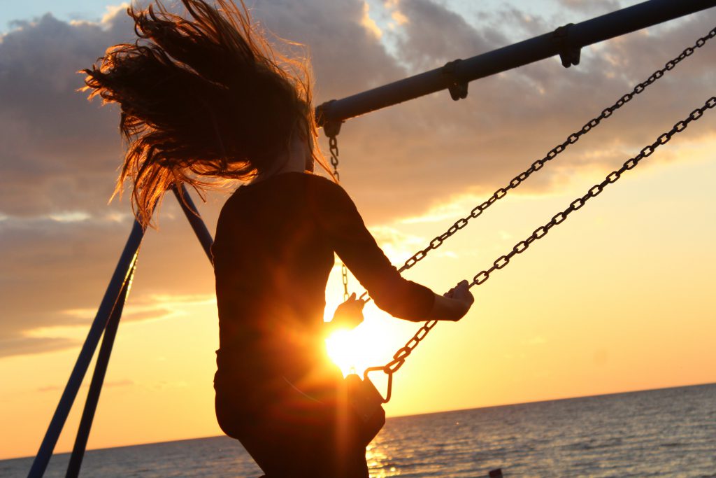 A young woman with free-flowing long hair swings on a metal play set next to the ocean. The sun is setting beyond her and the light catches in front of her lap as the swing descends. Here we look at five ways to start relying on God's promises.