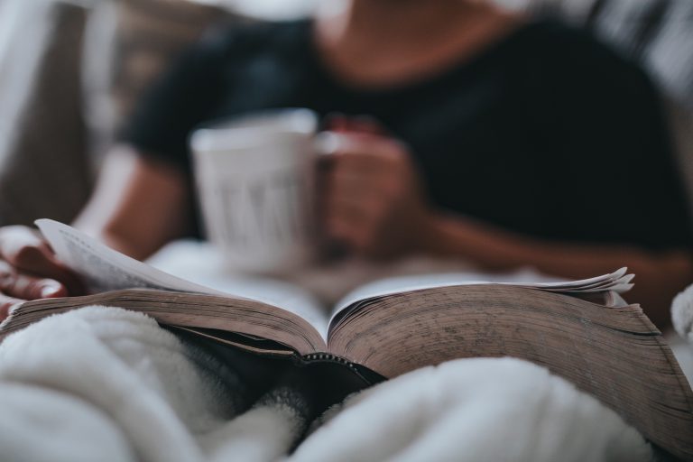 A person, of whom we can only see their hands holding a mug and thumbing through their Bible, and their shoulders as they sit in bed with a comfy white blanket covering their legs. The Bible is in focus, with the person out of focus in the near distance. Dive into your Bible study when you start with the gospel of John.